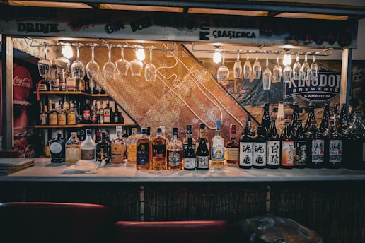A bar counter with a wide variety of bottles lined up, including whiskey and sake. Above the counter, several glasses hang upside down from a rack, and there are bright lights illuminating the area. Shelves in the background display more bottles, and there are signs featuring brands like Coca-Cola and Kingdom Brewery.