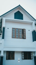 A modern two-story house with white walls and dark blue accents. The structure features multiple windows with wooden frames and grills, some of which are open. The design is minimalist with clean lines, and the house is illuminated by exterior lights.