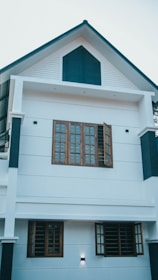 A modern two-story house with white walls and dark blue accents. The structure features multiple windows with wooden frames and grills, some of which are open. The design is minimalist with clean lines, and the house is illuminated by exterior lights.