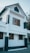 A modern two-story house with a gabled roof and a mix of white and dark gray exterior colors. Wooden framed windows provide a warm contrast to the clean lines of the structure. The house has a contemporary design with minimalistic features and an outside wall light fixture.
