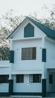 A modern two-story house with a white facade and dark accents around the windows and edges. The upper story features a prominent triangular gable with dark paneling. Multiple rectangular windows with wooden frames are visible on both floors. The roof is pitched and covered with dark shingles. Trees with sparse foliage are seen in the background, suggesting a natural setting.