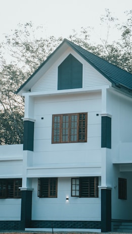 A modern two-story house with a white facade and dark accents around the windows and edges. The upper story features a prominent triangular gable with dark paneling. Multiple rectangular windows with wooden frames are visible on both floors. The roof is pitched and covered with dark shingles. Trees with sparse foliage are seen in the background, suggesting a natural setting.