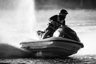 A person rides a jet ski at high speed across a body of water, creating a large spray. The individual is wearing a helmet and protective gear. The image is in black and white, adding a dramatic effect.