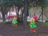 Colorful Christmas lights decorate a park setting. Illuminated figures resembling elves stand next to trees wrapped in string lights. Sparse leaves cover the ground, while a backdrop of trees and a building create a festive atmosphere.