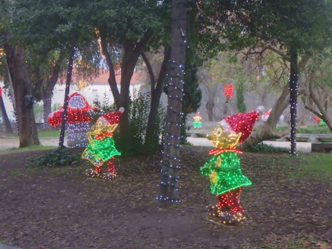 Colorful Christmas lights decorate a park setting. Illuminated figures resembling elves stand next to trees wrapped in string lights. Sparse leaves cover the ground, while a backdrop of trees and a building create a festive atmosphere.