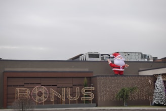 A large inflatable Santa Claus is positioned on the roof of a building. The facade features a sign that reads 'BONUS' illuminated with lights. There are two decorated Christmas trees on either side, adding to the festive theme.