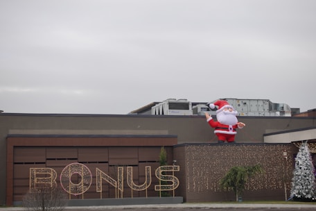 A large inflatable Santa Claus is positioned on the roof of a building. The facade features a sign that reads 'BONUS' illuminated with lights. There are two decorated Christmas trees on either side, adding to the festive theme.