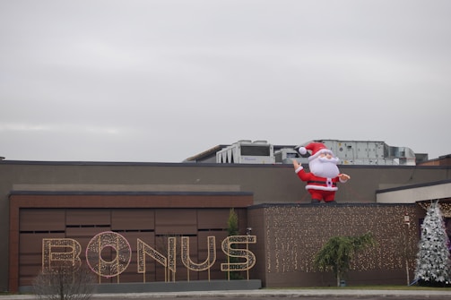 A large inflatable Santa Claus is positioned on the roof of a building. The facade features a sign that reads 'BONUS' illuminated with lights. There are two decorated Christmas trees on either side, adding to the festive theme.
