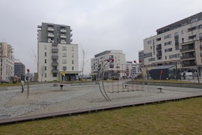 A modern residential area featuring several multi-story apartment buildings. In the foreground, there is a small playground with metal climbing structures and seesaws. The ground is covered with gravel and surrounded by a green grassy area. The environment appears to be calm with no people present.