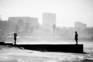 A black and white photograph depicts a person standing on a pier, looking towards another individual taking a photo of them. The city skyline is visible in the hazy background, with palm trees and buildings creating an urban coastal scene. The beach and waves appear in the foreground.