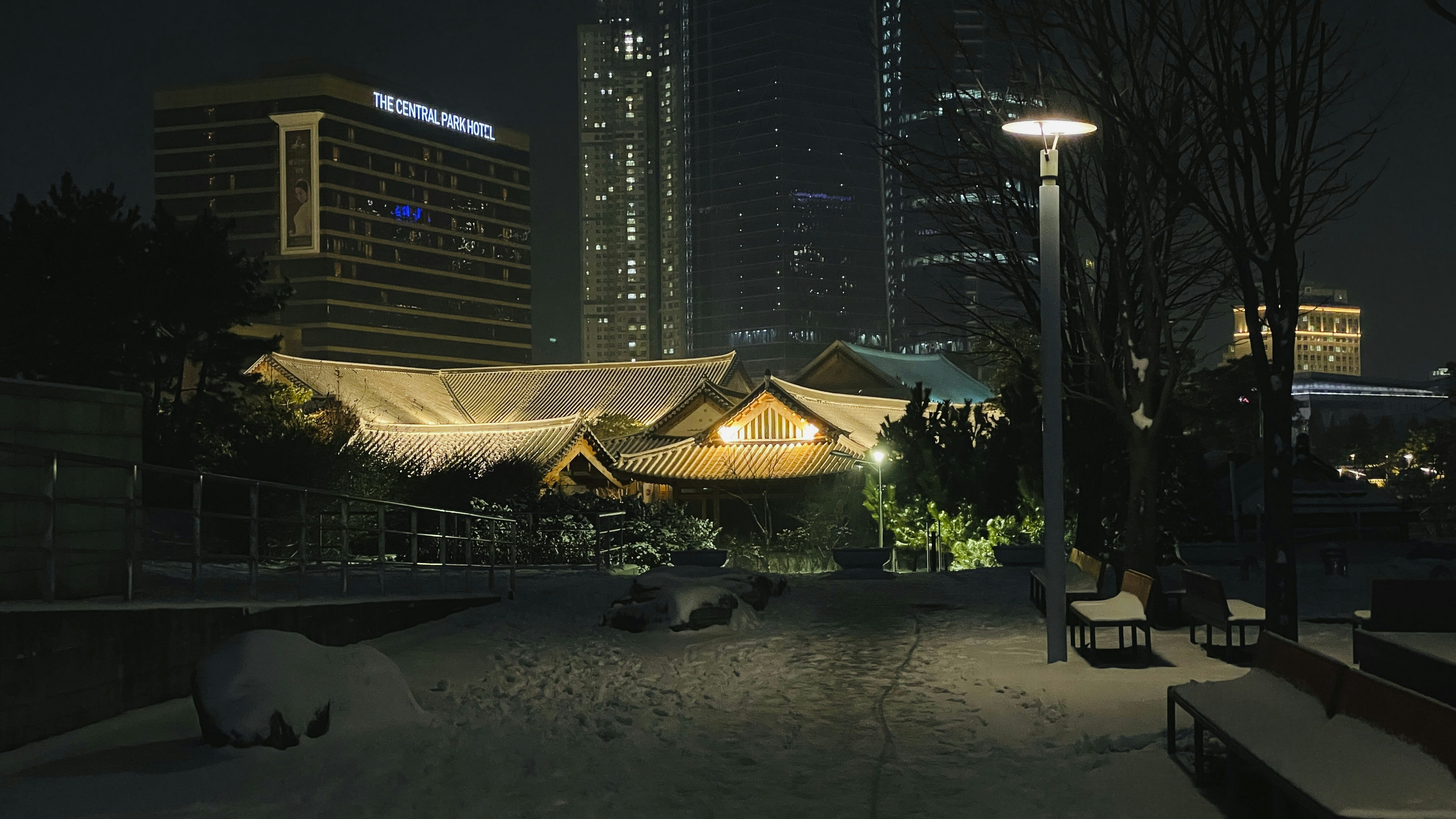 a snow covered park with benches and buildings in the background