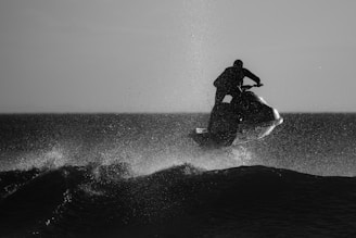 A person riding a jet ski is performing a jump over the crest of a wave, with water splashing around in a silhouette view. The light contrasts sharply with the figures creating an impressive action scene against the open water.