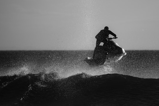 A person riding a jet ski is performing a jump over the crest of a wave, with water splashing around in a silhouette view. The light contrasts sharply with the figures creating an impressive action scene against the open water.