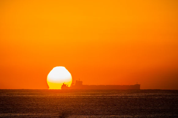 A large cargo ship at sea with crew members working on deck against a sunset backdrop.