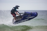A person riding a jet ski is captured mid-air over water. The individual is wearing a helmet and protective gear, and the jet ski is predominantly blue. Water is splashing around the vehicle as it skims through the waves, suggesting fast movement and excitement.
