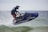 A person riding a jet ski is captured mid-air over water. The individual is wearing a helmet and protective gear, and the jet ski is predominantly blue. Water is splashing around the vehicle as it skims through the waves, suggesting fast movement and excitement.