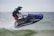 A person riding a jet ski is captured mid-air over water. The individual is wearing a helmet and protective gear, and the jet ski is predominantly blue. Water is splashing around the vehicle as it skims through the waves, suggesting fast movement and excitement.