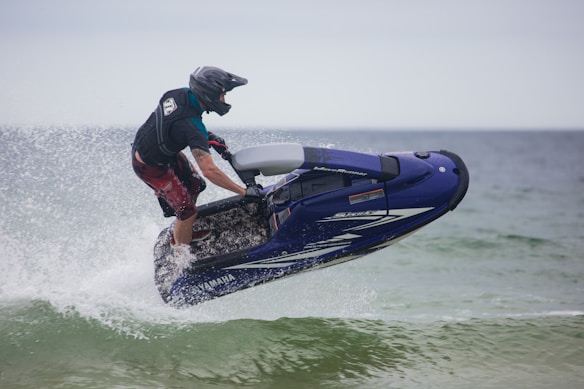 A person riding a jet ski is captured mid-air over water. The individual is wearing a helmet and protective gear, and the jet ski is predominantly blue. Water is splashing around the vehicle as it skims through the waves, suggesting fast movement and excitement.