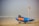 A rescue boat is positioned on a beach, attached to a trailer. The boat is blue with the word 'RESCUE' in large yellow and red letters. Next to the boat, a pole with a red and yellow flag is planted in the sand. The background shows a foggy view of the ocean.