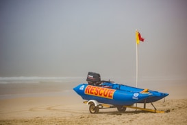 A rescue boat is positioned on a beach, attached to a trailer. The boat is blue with the word 'RESCUE' in large yellow and red letters. Next to the boat, a pole with a red and yellow flag is planted in the sand. The background shows a foggy view of the ocean.