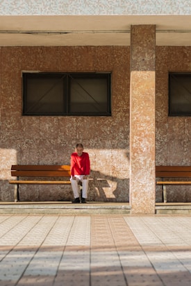 An elderly person wearing a red sweater and white pants is sitting on a wooden bench. The setting is outdoors, with two rectangular windows on a patterned brown wall behind the bench. The sunlight casts a shadow of the person and the bench on the floor.