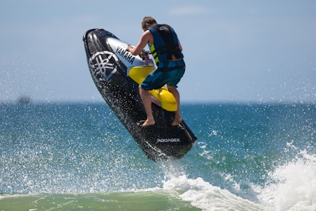 A person is riding a Yamaha jet ski, leaping out of the ocean. The backdrop features a vast expanse of blue sea and sky. The wave spray surrounds the vehicle, capturing the dynamic motion.