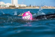 A swimmer in a body of water is wearing a bright pink swim cap with a cartoonish face and large white teeth design. The swimmer is in mid-stroke, splashing water around, with the cityscape and buildings visible in the distant background.