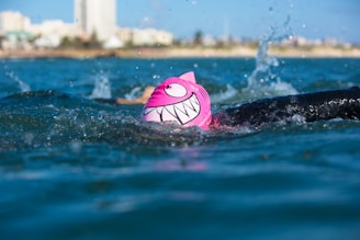 A swimmer in a body of water is wearing a bright pink swim cap with a cartoonish face and large white teeth design. The swimmer is in mid-stroke, splashing water around, with the cityscape and buildings visible in the distant background.