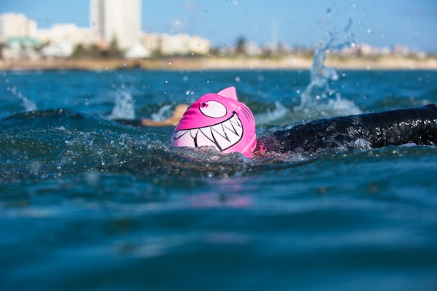 A swimmer in a body of water is wearing a bright pink swim cap with a cartoonish face and large white teeth design. The swimmer is in mid-stroke, splashing water around, with the cityscape and buildings visible in the distant background.