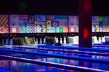 A bowling alley with several brightly lit lanes and pins arranged at the end. The back wall is adorned with vibrant, colorful abstract artwork, depicting various geometric shapes and playful figures. The alley is illuminated with blue and purple neon lights, adding a lively atmosphere.