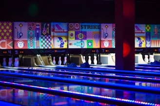 A bowling alley with several brightly lit lanes and pins arranged at the end. The back wall is adorned with vibrant, colorful abstract artwork, depicting various geometric shapes and playful figures. The alley is illuminated with blue and purple neon lights, adding a lively atmosphere.