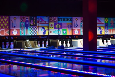 A bowling alley with several brightly lit lanes and pins arranged at the end. The back wall is adorned with vibrant, colorful abstract artwork, depicting various geometric shapes and playful figures. The alley is illuminated with blue and purple neon lights, adding a lively atmosphere.