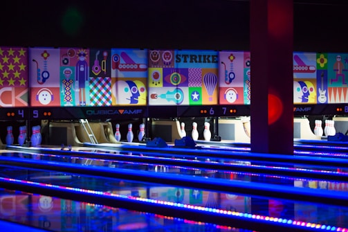 A bowling alley with several brightly lit lanes and pins arranged at the end. The back wall is adorned with vibrant, colorful abstract artwork, depicting various geometric shapes and playful figures. The alley is illuminated with blue and purple neon lights, adding a lively atmosphere.