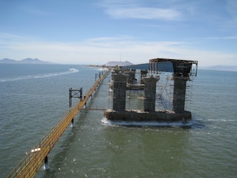 A partially constructed bridge extends over a body of water. The structure is supported by several large concrete pillars and metal scaffolding. In the distance, mountains are visible under a clear blue sky, and a boat creates a wake on the water's surface.