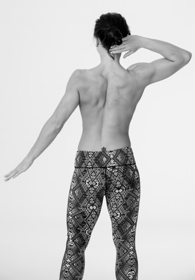 A person is seen from behind, with a muscular and toned back, stretching one arm up and the other extended to the side. They are wearing patterned leggings with geometric designs, and the image is in black and white.