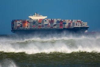 A large cargo ship filled with multi-colored shipping containers sailing through rough waters, with sprays of ocean waves visible in the foreground against a backdrop of a clear blue sky.