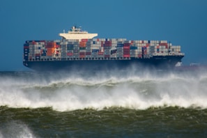 A large cargo ship filled with multi-colored shipping containers sailing through rough waters, with sprays of ocean waves visible in the foreground against a backdrop of a clear blue sky.