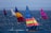 Colorful sailboats are cruising across a body of water with vibrant sails in various combinations of blue, red, yellow, and orange. The sunlight reflects off the waves, creating a sparkling effect on the water. The scene suggests a calm day with gentle winds perfect for sailing.