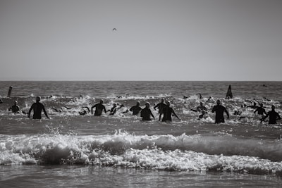 A group of people wearing wetsuits swim in the ocean, possibly participating in an open water race or event. The water is choppy with visible waves, and a distant horizon is visible under a clear sky.
