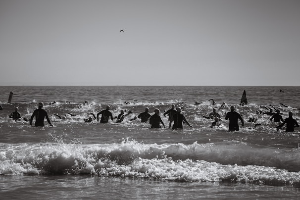 A group of people wearing wetsuits swim in the ocean, possibly participating in an open water race or event. The water is choppy with visible waves, and a distant horizon is visible under a clear sky.