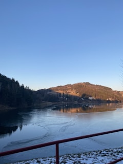 A serene frozen lake with surrounding hills and a clear blue sky. The lake's surface shows patches of ice and a few ducks. In the background, hills are illuminated by the late afternoon sun, and some scattered buildings are visible. A red railing is in the foreground, with a small amount of snow on the ground.