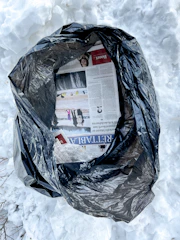 A black plastic trash bag is partially open, revealing torn pages of a newspaper and crumpled transparent plastic. The bag is placed on a surface covered with snow, suggesting a cold outdoor environment.