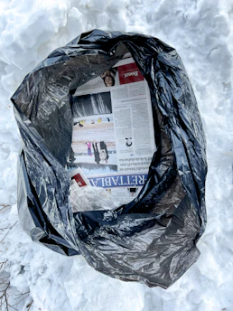 A black plastic trash bag is partially open, revealing torn pages of a newspaper and crumpled transparent plastic. The bag is placed on a surface covered with snow, suggesting a cold outdoor environment.