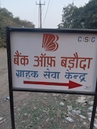 A sign with text written in Hindi, featuring a bank logo and an arrow. The sign is placed outdoors on a grassy area with some litter visible on the ground. In the background, there are trees and electrical poles with power lines stretching into the distance.
