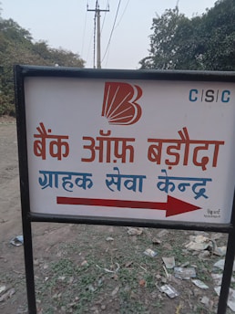 A sign with text written in Hindi, featuring a bank logo and an arrow. The sign is placed outdoors on a grassy area with some litter visible on the ground. In the background, there are trees and electrical poles with power lines stretching into the distance.