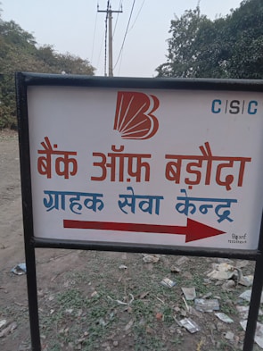 A sign with text written in Hindi, featuring a bank logo and an arrow. The sign is placed outdoors on a grassy area with some litter visible on the ground. In the background, there are trees and electrical poles with power lines stretching into the distance.