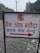 A sign with text written in Hindi, featuring a bank logo and an arrow. The sign is placed outdoors on a grassy area with some litter visible on the ground. In the background, there are trees and electrical poles with power lines stretching into the distance.