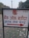 A sign with text written in Hindi, featuring a bank logo and an arrow. The sign is placed outdoors on a grassy area with some litter visible on the ground. In the background, there are trees and electrical poles with power lines stretching into the distance.