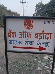 A sign with text written in Hindi, featuring a bank logo and an arrow. The sign is placed outdoors on a grassy area with some litter visible on the ground. In the background, there are trees and electrical poles with power lines stretching into the distance.