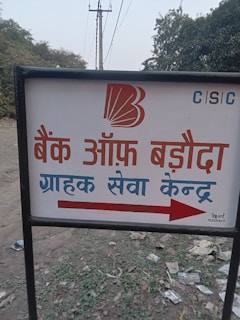 A sign with text written in Hindi, featuring a bank logo and an arrow. The sign is placed outdoors on a grassy area with some litter visible on the ground. In the background, there are trees and electrical poles with power lines stretching into the distance.